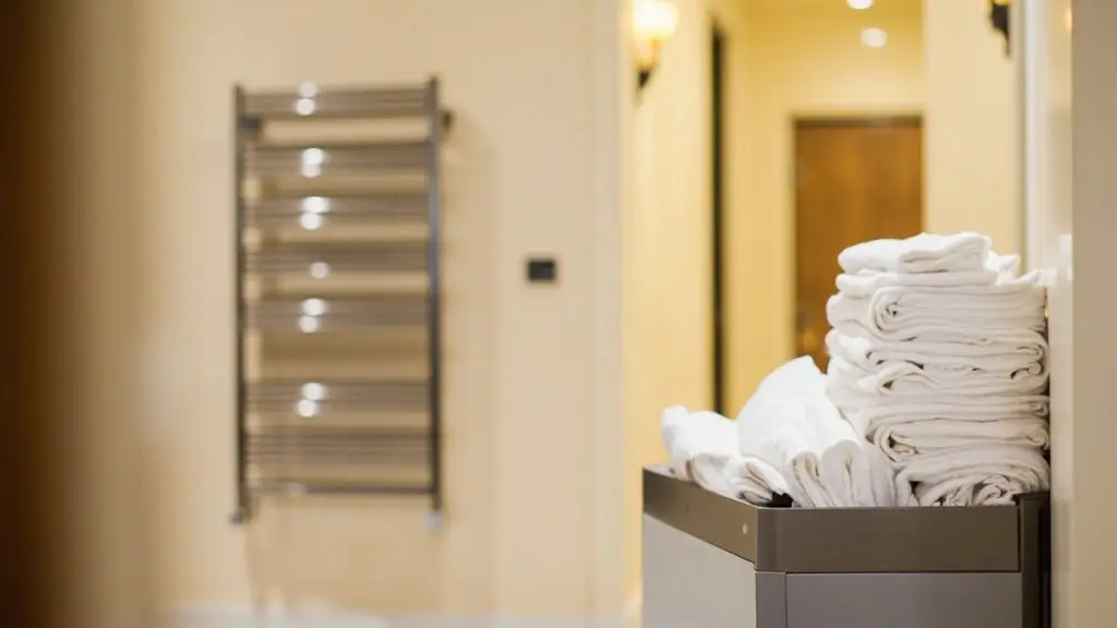 Hotel housekeeping linen cart with stacked white towels and bed linens in service corridor with heated towel rack on wall, commercial heated towel rack and towel warmer